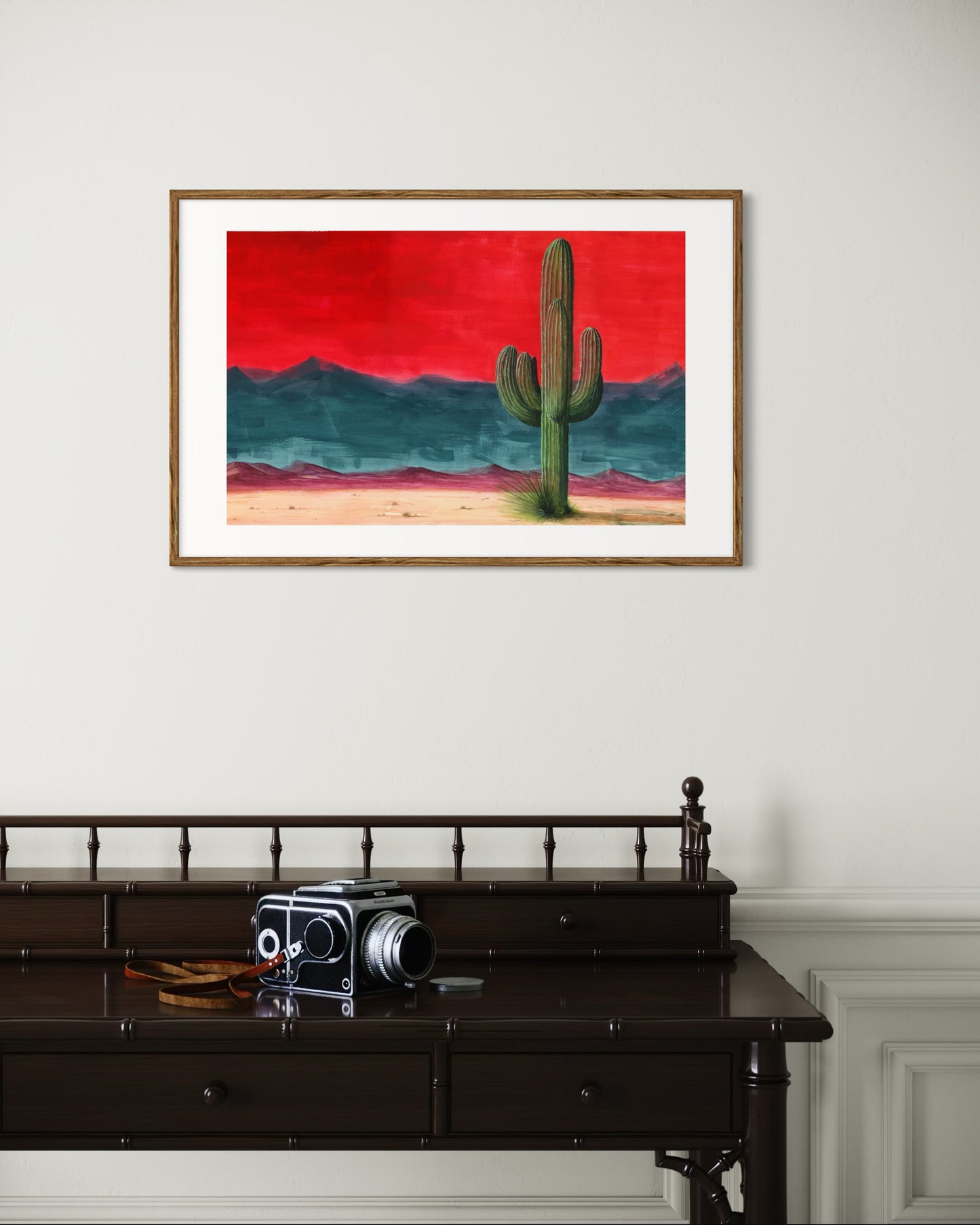 Framed artwork of a cactus against a red sky on a white wall above a dark wooden desk with a vintage camera.