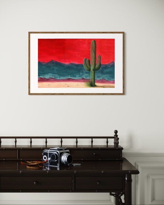 Framed artwork of a cactus against a red sky on a white wall above a dark wooden desk with a vintage camera.