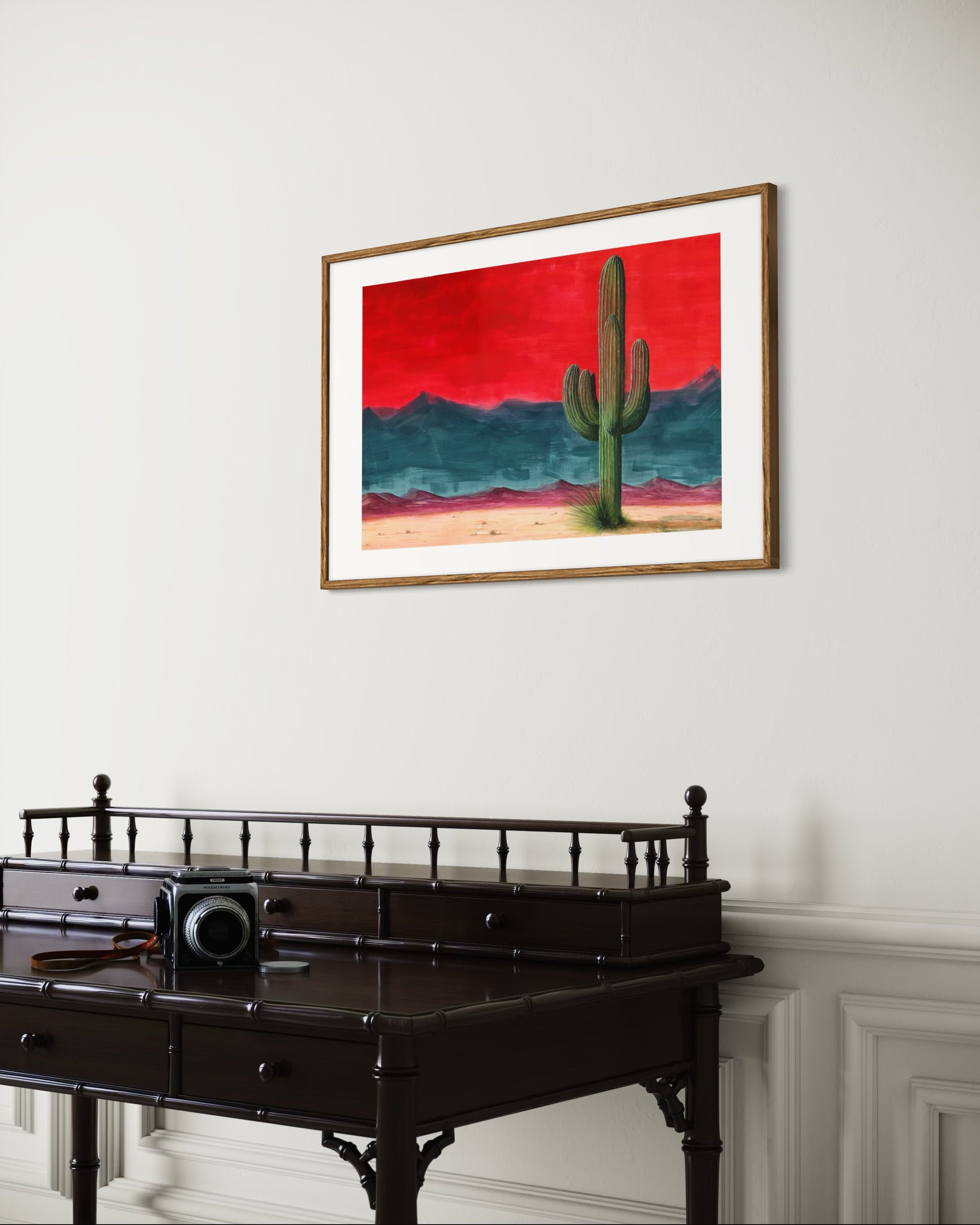Framed artwork of a cactus against a red sky on a white wall above a dark wooden desk.